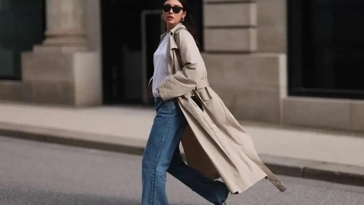 Mujer con gabardina beige, jeans acampanados y gafas de sol camina por la calle. ¡Estilo perfecto para la primavera 2026!