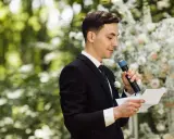 El maestro de ceremonias, con traje negro y micrófono, lee un discurso frente a un arco de flores.