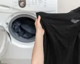 Manos sostienen ropa negra con manchas blancas, cerca de una lavadora abierta. ¿Por qué la ropa negra sale con manchas blancas?