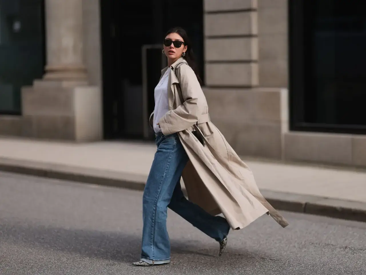 Mujer con gabardina beige, jeans acampanados y gafas de sol camina por la calle. ¡Estilo perfecto para la primavera 2026!