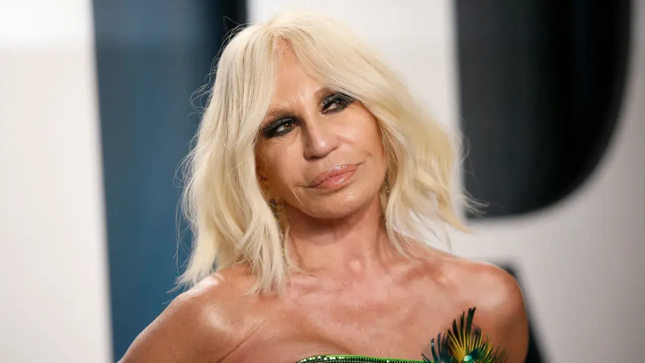Donatella Versace joven, con su icónico cabello rubio y maquillaje dramático, luce un vestido verde con detalles de plumas.