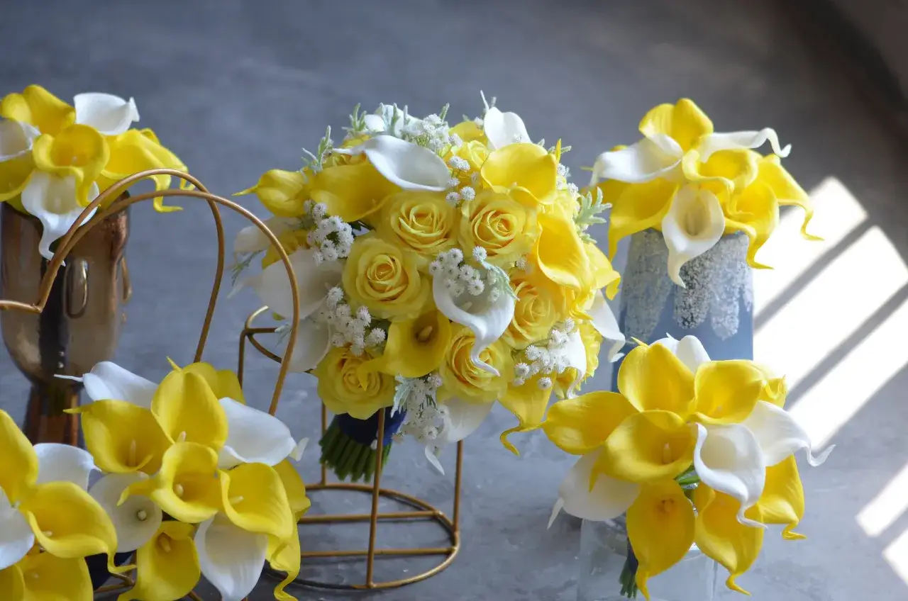 Arreglos florales con rosas amarillas y calas blancas. Un ramo principal destaca, rodeado por otros arreglos que irradian alegría.