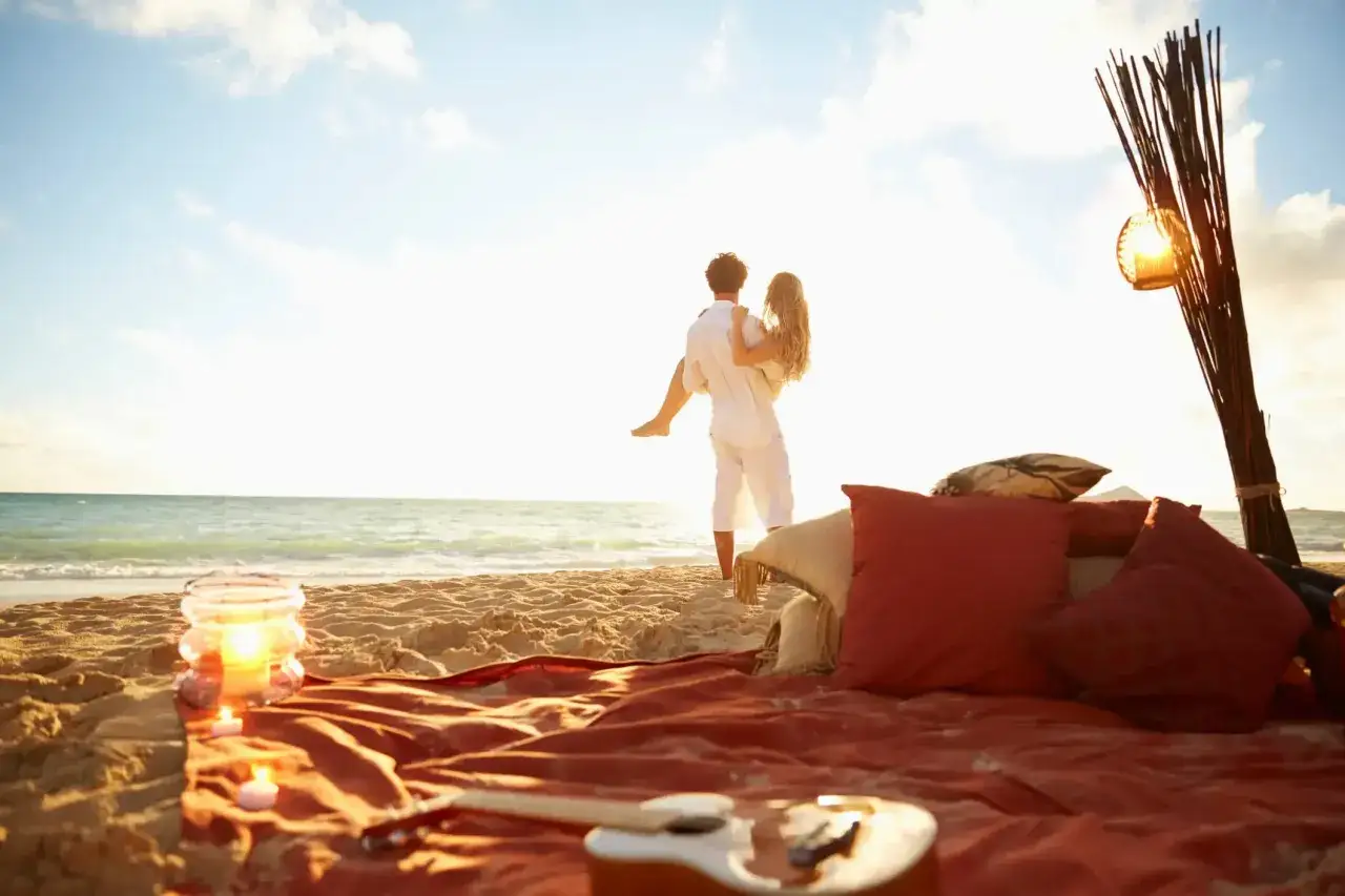 Pareja en la playa al atardecer, él la carga en brazos. Un rincón romántico para su luna de miel con cojines, velas y una guitarra.