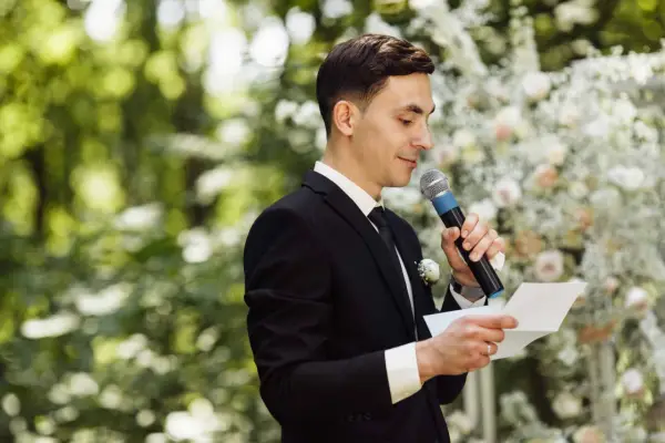 El maestro de ceremonias, con traje negro y micrófono, lee un discurso frente a un arco de flores.