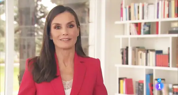 Reina Letizia Ortiz, sonriendo, con chaqueta roja y blusa de encaje blanco, frente a una estantería llena de libros.