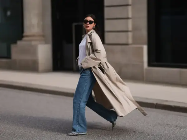 Mujer con gabardina beige, jeans acampanados y gafas de sol camina por la calle. ¡Estilo perfecto para la primavera 2026!