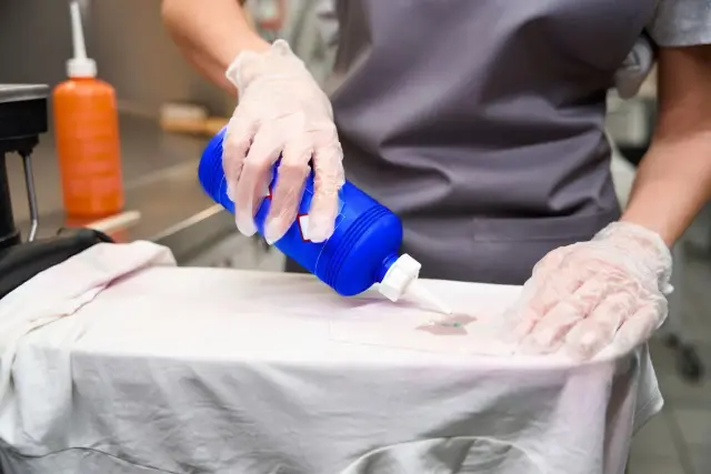 Manos enguantadas aplican líquido de una botella azul sobre una camiseta blanca, tratando manchas de prendas que destiñen en seco.