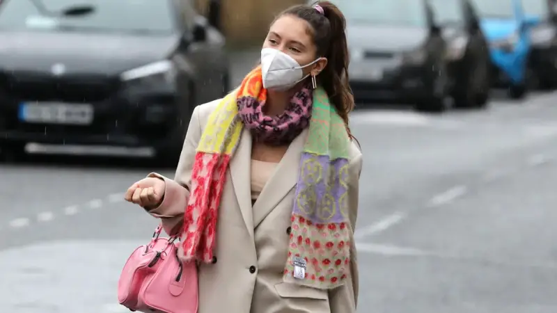 Iris Law, con mascarilla y bufanda multicolor, camina por la calle con un bolso rosa.