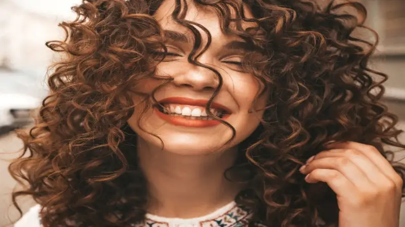 Mujer sonriendo con unos cortes de pelo rizado espectaculares, su cabello castaño cae en cascada alrededor de su rostro.